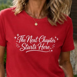 以下が含まれることがあります： 胸元に「The Next Chapter Starts Here」という白い文字と、小さな星や線のグラフィックがプリントされた赤い半袖Tシャツ。クルーネックで、茶色のレザーベルトと白いパンツを合わせて着用しています。