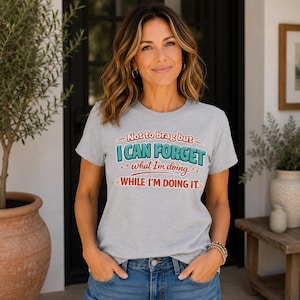 May include: A light gray short-sleeved t-shirt featuring the text Not to brag but I CAN FORGET what I'm doing WHILE I'M DOING IT. The graphic design uses a combination of red and teal lettering with small star accents printed on the front chest.