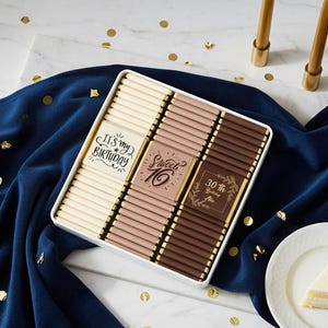 May include: A white box filled with chocolate bars, with the text "It's my Birthday", "Sweet 16", and "30th Birthday". Two gold candles and a slice of cake are also present. The scene is set on a marble surface with a blue cloth and gold confetti.