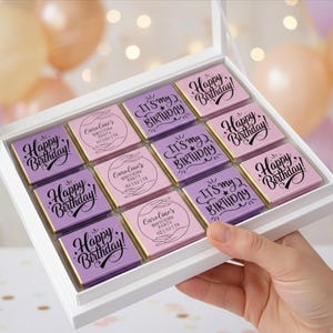 Peut inclure: Une boîte blanche avec un couvercle transparent contenant douze carrés de chocolat. Les carrés sont violets et roses, avec des bords dorés et des inscriptions d'anniversaire, comme "Happy Birthday!"
