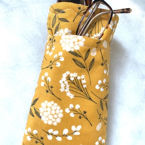 May include: A mustard yellow fabric glasses case featuring a white floral and leaf pattern. The case is quilted with visible stitching along the top edge and contains a pair of brown tortoiseshell eyeglasses tucked inside the opening.