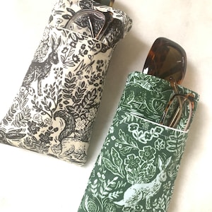 May include: Two fabric eyeglass cases with a woodland print. One case is green with white rabbits and the other is cream with black rabbits. Both cases have a pocket for glasses.