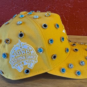May include: A bright yellow baseball cap decorated with multiple jewel-like studs. The cap has a patch with the text "1986 ALL STAR GAME". The jewels are in various colours, including blue, black, and clear, adding a touch of sparkle.