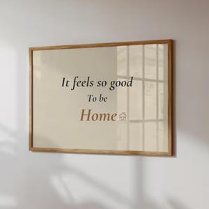 May include: A framed wall art piece with the text "It feels so good to be Home" in black and brown font. The artwork has a light beige background and a wooden frame. A small house icon is next to the word "Home".