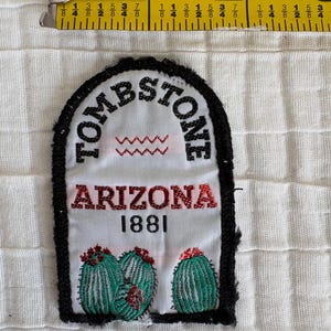 May include: A rectangular patch with the word "TOMBSTONE" arched across the top in black letters. Below, the word "ARIZONA" is in red, with "1881" beneath it. The patch features embroidered green cacti with red flowers. A black border surrounds the white background.