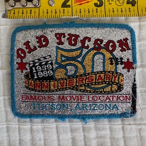 May include: A rectangular patch with the text "OLD TUCSON 50TH ANNIVERSARY" in red and gold. The patch includes the years 1939 and 1989, and the words "FAMOUS MOVIE LOCATION TUCSON ARIZONA". The patch is approximately 3 inches wide.