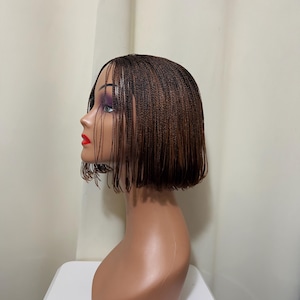 May include: A brown braided wig styled in a short bob, displayed on a mannequin head. The wig features a side part and fine braids. The mannequin has red lipstick and purple eyeshadow. The background is a plain white wall.