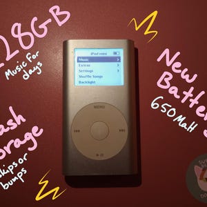 May include: A silver iPod mini digital music player with a circular click wheel and a screen displaying a menu with options for Music, Extras, Settings, Shuffle Songs, and Backlight. Text on the image includes 128GB, Music for days, New Battery 650mAh, Flash Storage, and No skips or bumps.