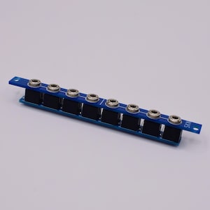 May include: A blue circuit board featuring eight audio jacks. Each jack is silver and black. The jacks are arranged in a row along the board. The board is rectangular with a small extension. The text "SKII" is printed on the board.