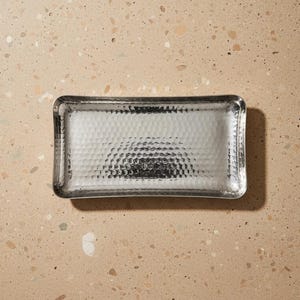 May include: A rectangular, clear glass tray with a hammered metal interior. The tray has rounded corners and a textured surface. The tray is designed for decorative or functional use.