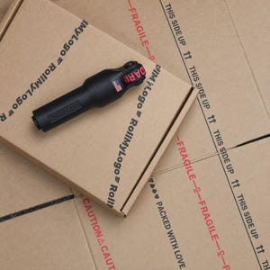 May include: A black cylindrical tool with a small American flag emblem and red text reading DARR on the top end. The tool features the embossed text RollMyLogo on its side and rests on a brown cardboard box printed with shipping labels.