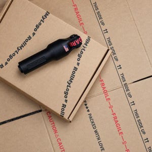 May include: A black cylindrical tool with a textured handle and a red strap featuring the word DARK in black letters. A small American flag emblem is visible on the side of the tool. The tool is resting on a brown cardboard box with printed text.