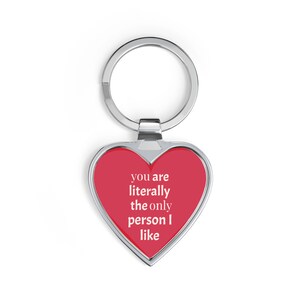 Könnte beinhalten: Herzförmiger Schlüsselanhänger mit roter Mitte und silberner Umrandung. Der Schlüsselanhänger trägt den weißen Text "you are literally the only person I like". Er ist an einem silbernen Schlüsselring befestigt.