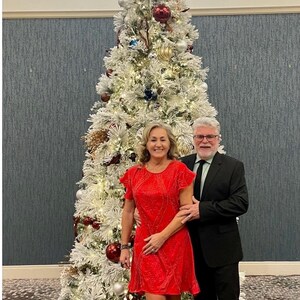 May include: A couple poses in front of a decorated Christmas tree. The tree is white with ornaments in red, silver, and gold. The woman wears a red dress, and the man wears a black suit. The tree is lit with white lights.