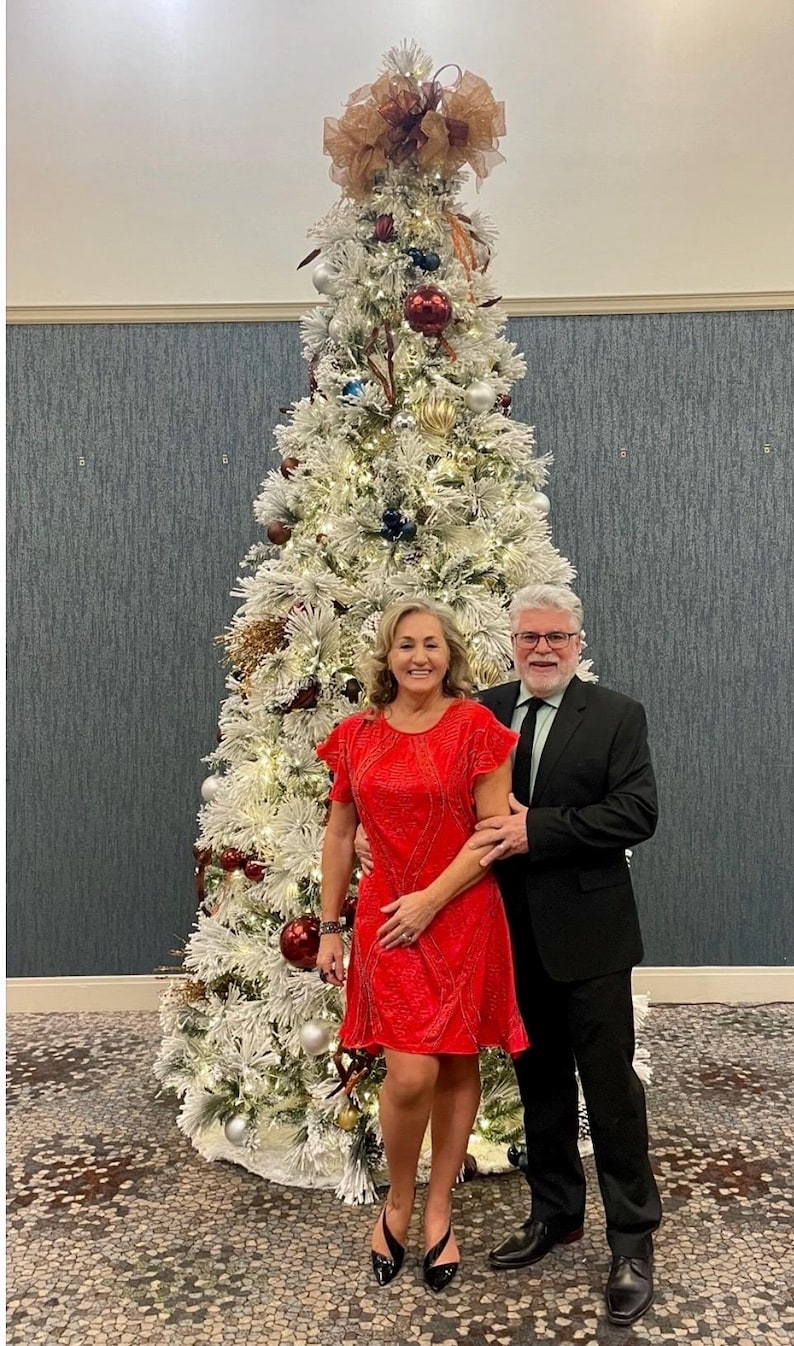 May include: A couple poses in front of a decorated Christmas tree. The tree is white with ornaments in red, silver, and gold. The woman wears a red dress, and the man wears a black suit. The tree is lit with white lights.