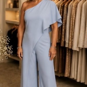 Op de afbeelding: Lichtblauwe jumpsuit met een off-shoulder ontwerp en wijde pijpen. De top heeft een gedrapeerd ruchedetail op de schouder. De jumpsuit is gemaakt van een gladde stof en wordt gedragen door een persoon in een kledingwinkel.