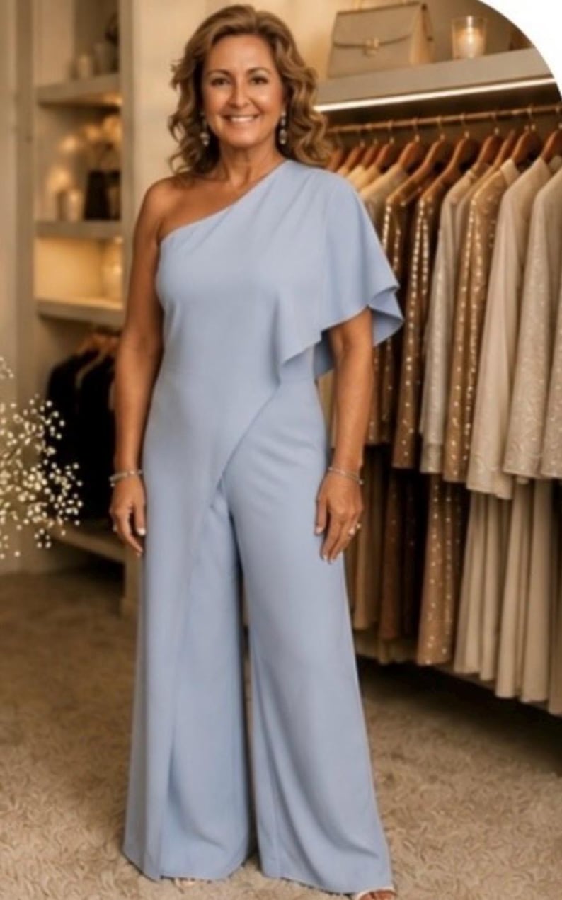 Op de afbeelding: Lichtblauwe jumpsuit met een off-shoulder ontwerp en wijde pijpen. De top heeft een gedrapeerd ruchedetail op de schouder. De jumpsuit is gemaakt van een gladde stof en wordt gedragen door een persoon in een kledingwinkel.