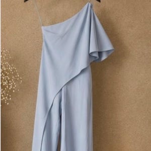Op de afbeelding: Een lichtblauwe jumpsuit met &eacute;&eacute;n schouder en een gedrapeerde overlay. De broek met wijde pijpen is zichtbaar en het kledingstuk hangt aan een zwarte hanger. De achtergrond is een getextureerde, lichtbruine kleur.