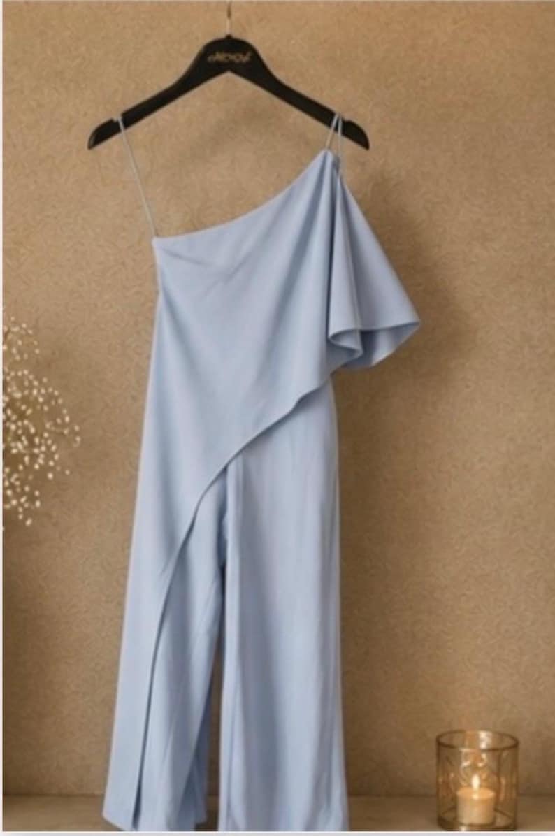 Op de afbeelding: Een lichtblauwe jumpsuit met &eacute;&eacute;n schouder en een gedrapeerde overlay. De broek met wijde pijpen is zichtbaar en het kledingstuk hangt aan een zwarte hanger. De achtergrond is een getextureerde, lichtbruine kleur.
