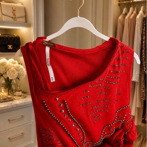 May include: A red sleeveless top with intricate beadwork and embellishments. The top is hanging on a white hanger in a well-lit closet. The design includes a scoop neckline and a unique asymmetrical cut.
