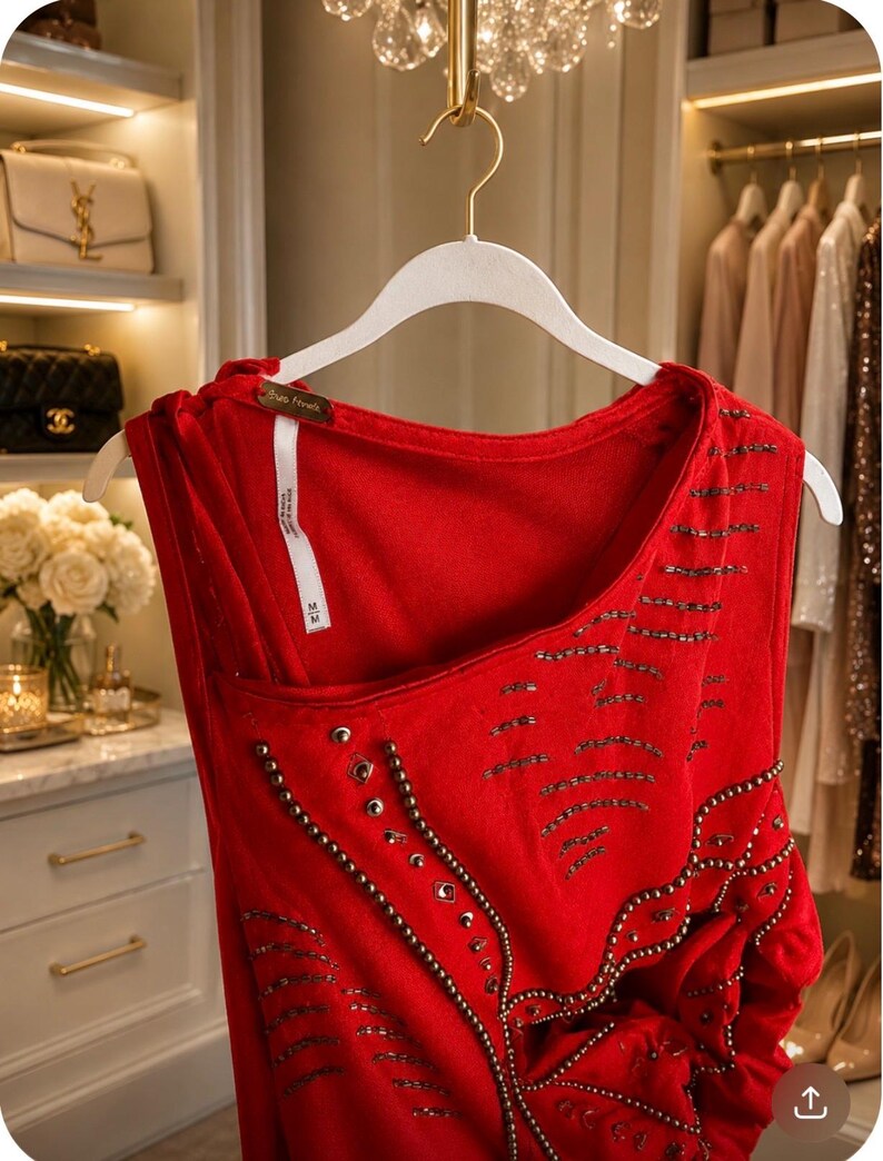 May include: A red sleeveless top with intricate beadwork and embellishments. The top is hanging on a white hanger in a well-lit closet. The design includes a scoop neckline and a unique asymmetrical cut.