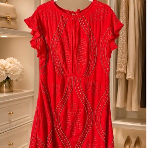 May include: A vibrant red dress with short, ruffled sleeves hangs on a white hanger. The dress features intricate beaded detailing and a flowing silhouette. The dress is displayed in a well-lit closet, suggesting a focus on fashion and apparel.