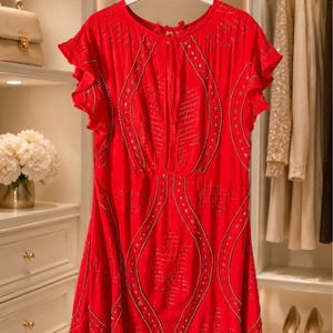 May include: A bright red dress with short, ruffled sleeves hangs on a white hanger. The dress features intricate silver-colored embellishments and a flowing silhouette. The background includes a closet with other clothing and accessories.