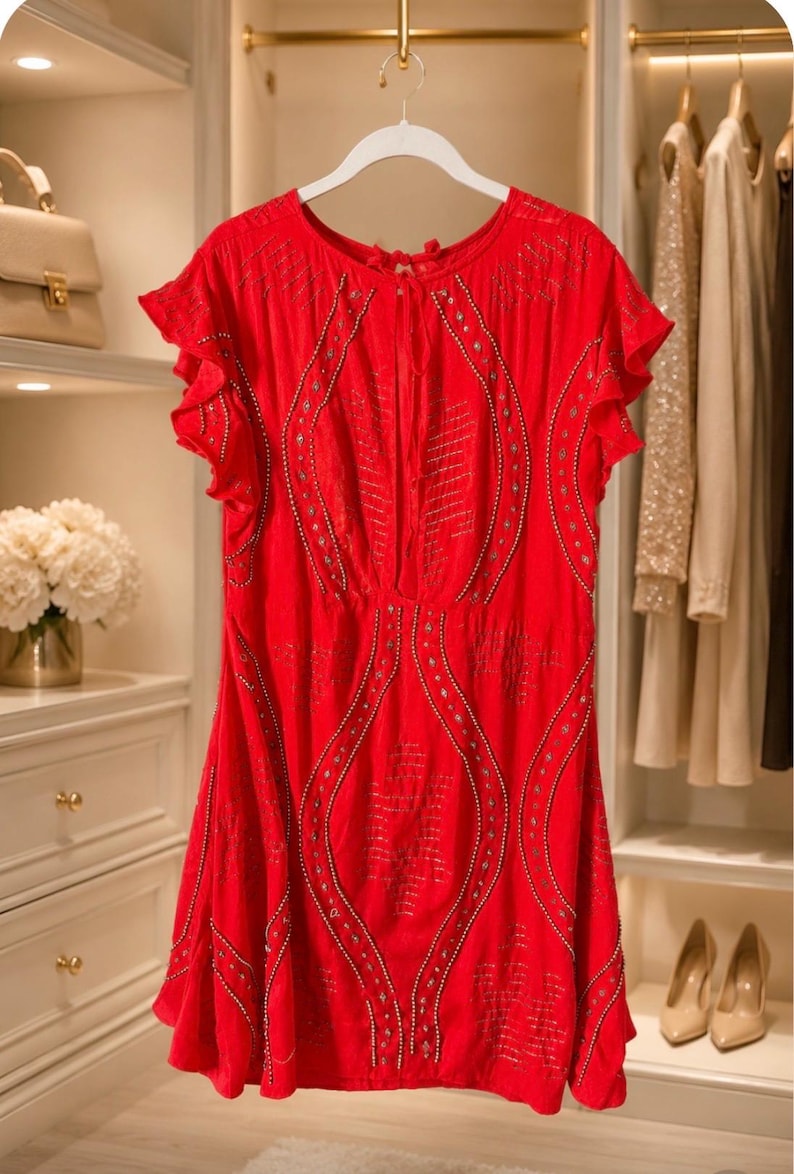 May include: A bright red dress with short, ruffled sleeves hangs on a white hanger. The dress features intricate silver-colored embellishments and a flowing silhouette. The background includes a closet with other clothing and accessories.