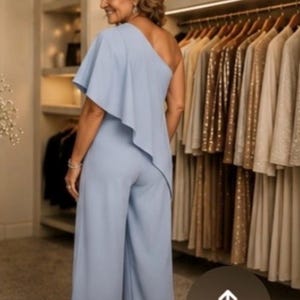 Op de afbeelding: Een lichtblauwe jumpsuit met &eacute;&eacute;n schouder en een vloeiend ruchedetail. De broek met wijde pijpen is zichtbaar. De achtergrond toont een kledingkast met kledingrekken en planken.