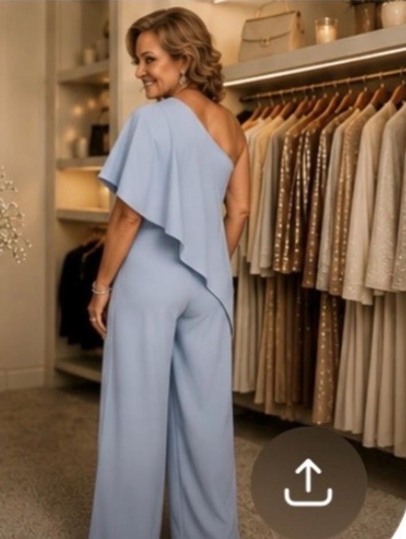 Op de afbeelding: Een lichtblauwe jumpsuit met &eacute;&eacute;n schouder en een vloeiend ruchedetail. De broek met wijde pijpen is zichtbaar. De achtergrond toont een kledingkast met kledingrekken en planken.
