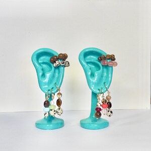 May include: Two turquoise ear-shaped jewelry displays holding multiple earrings and ear cuffs. The jewelry features various round beads in brown, white with black spots, pink, red, and translucent tones, all suspended from gold-toned metal hoops.