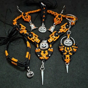 Può includere: Un set di gioielli in macramè con cordino nero e arancione, composto da una collana con ciondolo a forma di zucca argentata e pendente a punta, un bracciale con ciondolo a zucca e orecchini con ciondoli a forma di fantasma e zucca.