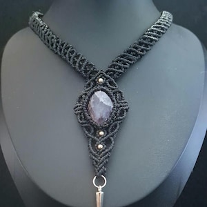 Può includere: Una collana in macramè nero con una pietra viola ovale centrale incastonata in un complesso lavoro a nodi. Il design include piccole perline metalliche e un ciondolo a punta color argento appeso a un anello circolare alla base.