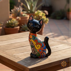 May include: A black ceramic cat figurine with a colorful, patterned body. The cat has a glossy finish and is sitting on a wooden table. The body of the cat is decorated with orange, yellow, and green floral designs.