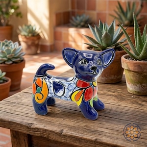 Puede incluir: Figura de chihuahua de cerámica en un vibrante esmalte azul con coloridos diseños florales y geométricos. El perro tiene orejas grandes y está posado sobre una superficie de madera, con plantas en macetas al fondo.
