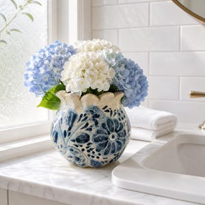 May include: A round ceramic vase with a scalloped rim, featuring a cream-colored base with intricate dark blue floral patterns. The vase is filled with a bouquet of fresh hydrangea flowers in shades of light blue and white, displaying full, rounded blooms.