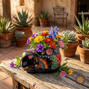 May include: A black ceramic turtle-shaped planter filled with colorful flowers, including purple, yellow, red, and blue blooms. The planter has a painted butterfly design and sits on a weathered wooden table. Other potted plants are in the background.
