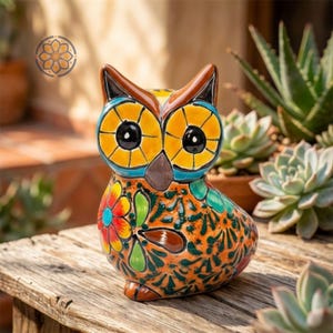 May include: A ceramic owl figurine with a colorful floral design. The owl has large yellow eyes, a brown beak, and a patterned orange body. The figurine is sitting on a wooden surface, with plants in the background.