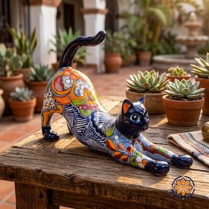 May include: A ceramic cat figurine in a stretching pose, featuring a vibrant, hand-painted design. The cat has a dark blue head and tail, with colorful floral and geometric patterns in orange, blue, and green across its body. It rests on a wooden table.