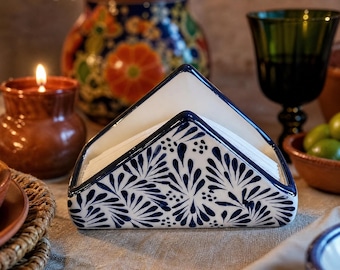 May include: A triangular ceramic napkin holder featuring a white base with a hand-painted dark blue floral and leaf pattern. The holder has a smooth, glossy finish and is designed to hold a stack of folded paper napkins in an upright position.