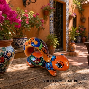May include: A hand-painted ceramic squirrel figurine featuring a glossy orange base color with intricate floral and geometric patterns in blue, green, red, and yellow. The squirrel is depicted in a curved, leaping pose with a large, arched tail.