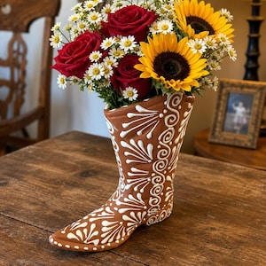 May include: A terracotta-colored ceramic vase shaped like a cowboy boot, decorated with white painted floral and swirl patterns. The vase is filled with a vibrant bouquet of deep red roses, bright yellow sunflowers, and small white daisies.
