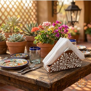 Puede incluir: Una mesa de madera rústica preparada para una comida al aire libre. Incluye un servilletero pintado a mano con diseños florales blancos, platos coloridos, vasos con bordes azules y plantas en macetas. La escena está iluminada por luz natural.