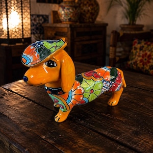 May include: A ceramic dachshund figurine featuring a glossy orange base color with vibrant, hand-painted floral patterns in shades of green, red, and blue. The dog wears a matching hat and a collar, with large dark eyes and a smooth, glazed finish.