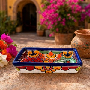 Puede incluir: Un plato rectangular de cerámica con un vibrante patrón pintado a mano en colores llamativos como azul, naranja, rojo, verde y amarillo. El plato tiene un borde azul grueso y sólido, una base blanca y motivos florales y geométricos complejos.