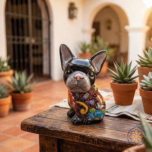 May include: A black ceramic French Bulldog figurine with a colorful floral design on its body. The dog has a pink nose and is sitting on a wooden table. The background includes potted plants and an outdoor setting.