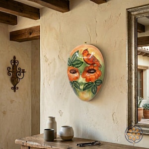 May include: Oval-shaped ceramic wall art featuring a red bird perched above orange flowers and green leaves. The artwork is painted on a cream-colored background and hangs on a textured wall. A decorative mirror and wooden table are also visible.