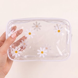 Peut inclure: Une trousse de maquillage rectangulaire transparente avec une fermeture éclair et une bordure blanches. Le sac est décoré de marguerites blanches avec des centres jaunes. Le sac est tenu par une main.