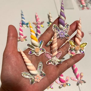 May include: A collection of colorful unicorn horn nail art decorations. The horns are various colors, including pink, yellow, and purple, with glitter and floral accents. The decorations are held in a hand, with several others visible in the background.