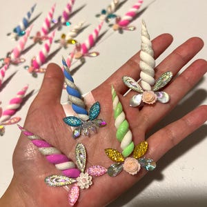 May include: A collection of colorful unicorn horn decorations. The horns are various colors, including pink, blue, green, and white, with glitter and floral accents. Some have a spiral design, and others are straight.